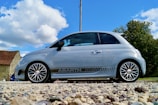 A small silver hatchback car with sporty detailing is parked on a gravel surface. The car features distinctive rims and branding on its side. In the background, there's a rustic stone building with a red roof, along with trees under a bright blue sky with fluffy white clouds.