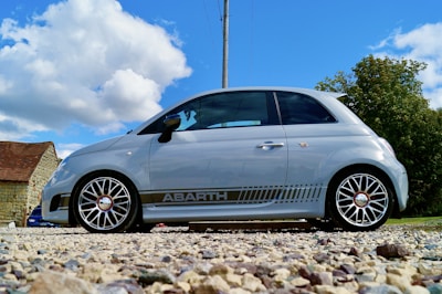 A small silver hatchback car with sporty detailing is parked on a gravel surface. The car features distinctive rims and branding on its side. In the background, there's a rustic stone building with a red roof, along with trees under a bright blue sky with fluffy white clouds.