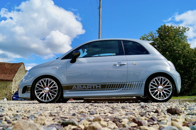A small silver hatchback car with sporty detailing is parked on a gravel surface. The car features distinctive rims and branding on its side. In the background, there's a rustic stone building with a red roof, along with trees under a bright blue sky with fluffy white clouds.