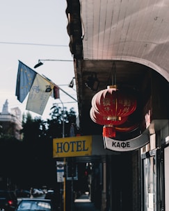 A street scene features bright red lanterns with Chinese characters hanging from a building. Nearby are signs including a yellow 'HOTEL' sign and a 'КАФЕ' sign written in Cyrillic letters. Flags with European Union symbols wave in the background, while a blue sky and some trees are visible.