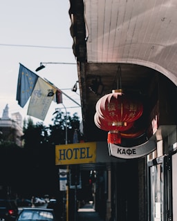 A street scene features bright red lanterns with Chinese characters hanging from a building. Nearby are signs including a yellow 'HOTEL' sign and a 'КАФЕ' sign written in Cyrillic letters. Flags with European Union symbols wave in the background, while a blue sky and some trees are visible.