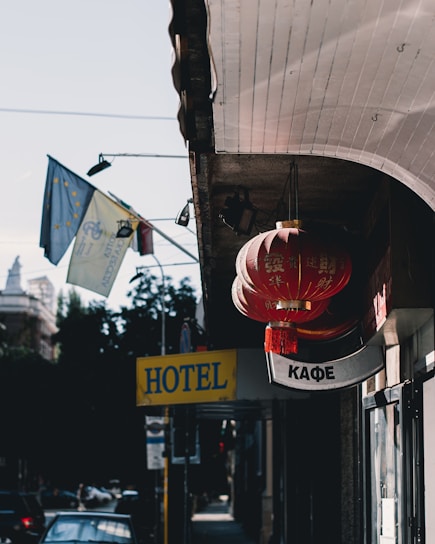 A street scene features bright red lanterns with Chinese characters hanging from a building. Nearby are signs including a yellow 'HOTEL' sign and a 'КАФЕ' sign written in Cyrillic letters. Flags with European Union symbols wave in the background, while a blue sky and some trees are visible.