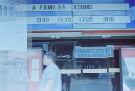 An outdoor cinema marquee displays showtimes for a movie titled 'A Familia Addams'. Below it, a blurry figure walks past, and reflections are visible on the glass doors in the background. The scene gives a slightly nostalgic, vintage film vibe.