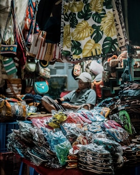 A market stall filled with a variety of hanging bags and tropical-themed textiles. The foreground features a table piled with stacks of packed shoes and sandals. In the background, a person is sitting, appearing relaxed and possibly occupied with a phone or book. The scene is dimly lit, giving it a cozy and slightly cluttered atmosphere.