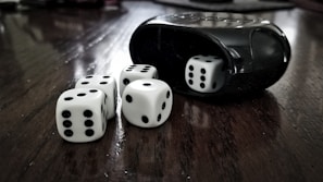 Five white dice with black dots are scattered on a dark wooden surface. A black dice cup is visible on the right, with one die partially inside it. The arrangement has a high contrast between the smooth texture of the dice and cup and the wood grain below.