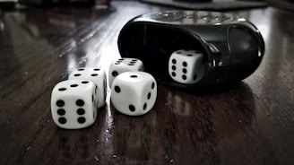 Five white dice with black dots are scattered on a dark wooden surface. A black dice cup is visible on the right, with one die partially inside it. The arrangement has a high contrast between the smooth texture of the dice and cup and the wood grain below.
