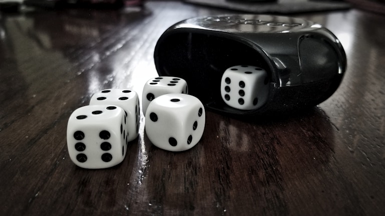 Five white dice with black dots are scattered on a dark wooden surface. A black dice cup is visible on the right, with one die partially inside it. The arrangement has a high contrast between the smooth texture of the dice and cup and the wood grain below.