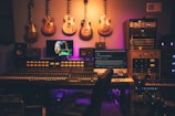 A music production studio featuring a large mixing console with numerous controls and sliders. Several electric guitars hang on the wall above the console. There are multiple monitors displaying digital audio software and a famous painting on one screen. The room is filled with various sound equipment, including speakers and audio modules. The lighting has a warm, ambient glow.