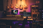 A music production studio featuring a large mixing console with numerous controls and sliders. Several electric guitars hang on the wall above the console. There are multiple monitors displaying digital audio software and a famous painting on one screen. The room is filled with various sound equipment, including speakers and audio modules. The lighting has a warm, ambient glow.