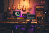 A music production studio featuring a large mixing console with numerous controls and sliders. Several electric guitars hang on the wall above the console. There are multiple monitors displaying digital audio software and a famous painting on one screen. The room is filled with various sound equipment, including speakers and audio modules. The lighting has a warm, ambient glow.