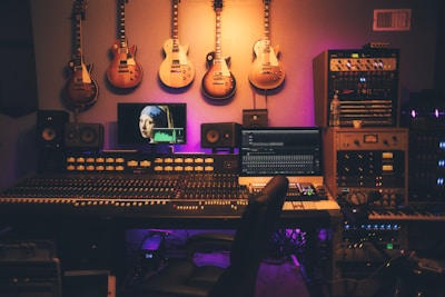 A music production studio featuring a large mixing console with numerous controls and sliders. Several electric guitars hang on the wall above the console. There are multiple monitors displaying digital audio software and a famous painting on one screen. The room is filled with various sound equipment, including speakers and audio modules. The lighting has a warm, ambient glow.