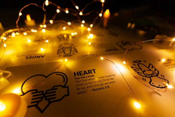 Fairy lights drape softly over a sheet of paper displaying religious symbols and text about love from Romans 5:8. The warm glow of the lights creates a peaceful and inviting atmosphere.