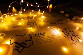 Fairy lights drape softly over a sheet of paper displaying religious symbols and text about love from Romans 5:8. The warm glow of the lights creates a peaceful and inviting atmosphere.