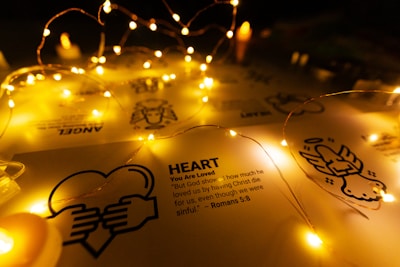 Fairy lights drape softly over a sheet of paper displaying religious symbols and text about love from Romans 5:8. The warm glow of the lights creates a peaceful and inviting atmosphere.