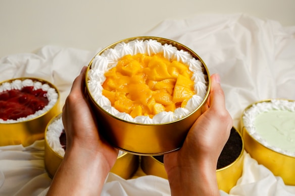 Hands holding a round cake with whipped cream around the edges and topped with pieces of mango, surrounded by other cakes with different toppings.