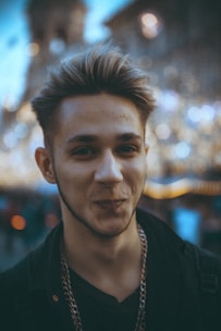 A young man with styled hair stands outdoors, wearing a dark outfit with a necklace. The background has a bokeh effect with blurred lights that suggest an evening setting.
