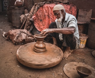 An elderly man is engaged in pottery, shaping a clay pot on a spinning wheel. He is seated on the ground in an outdoor, rustic setting, surrounded by various containers and materials. The scene conveys a sense of dedication and skill in traditional craftsmanship.