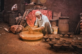 An elderly man is shaping clay on a traditional pottery wheel outside, surrounded by finished clay pots and rustic containers. The scene is set against a rough wall and amidst a variety of materials like burlap sacks and barrels.