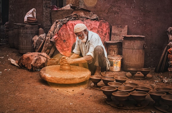 An elderly man is shaping clay on a traditional pottery wheel outside, surrounded by finished clay pots and rustic containers. The scene is set against a rough wall and amidst a variety of materials like burlap sacks and barrels.