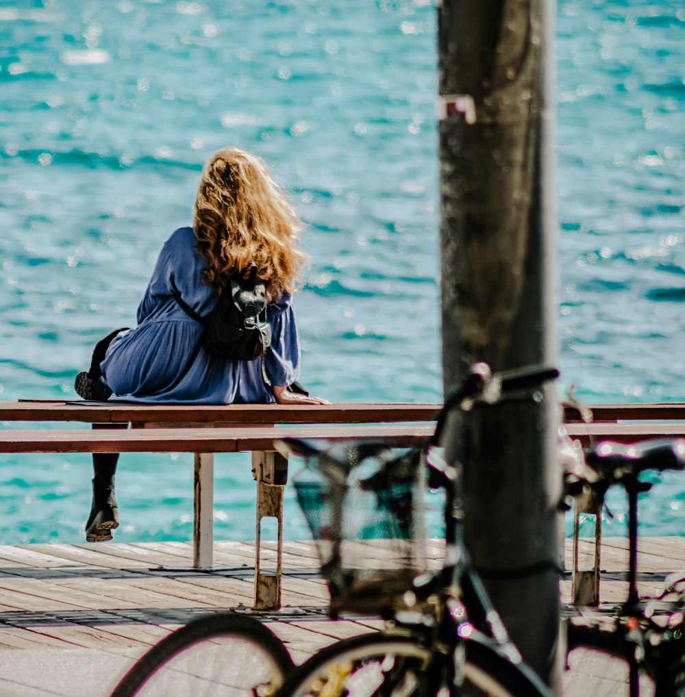 woman in black jacket sitting on brown wooden bench beside body of water during daytime