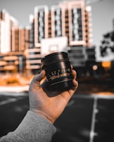 A hand holds a camera lens in the foreground, with a blurred urban landscape of tall buildings in the background. The scene is artistically framed with focus on the lens, while the buildings appear out of focus, emphasizing the photographic theme.