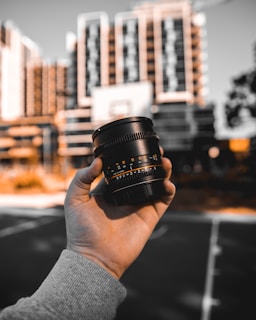 A hand holds a camera lens in the foreground, with a blurred urban landscape of tall buildings in the background. The scene is artistically framed with focus on the lens, while the buildings appear out of focus, emphasizing the photographic theme.