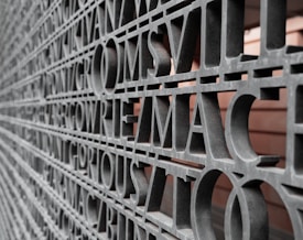 A close-up view of a wall featuring large, metallic letters arranged in a grid pattern. The letters are evenly spaced and appear to form words, creating a textured, sculptural effect. The background is visible through the spaces between the letters, adding depth to the composition.