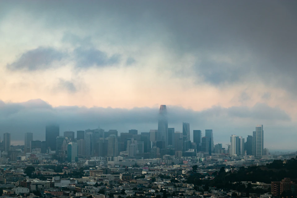 The image presents a panoramic view of the San Francisco Bay Area, with the iconic Golden Gate Bridge visible in the distance. The cityscape is densely populated with numerous buildings and skyscrapers, showcasing the urban landscape. The sky above is filled with clouds, casting a soft light over the scene. The perspective of the photo is from a high vantage point, providing an expansive view of the bay area and its surroundings.