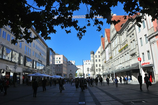 A bustling city street flanked by historic buildings with ornate facades, featuring numerous pedestrians and outdoor cafe seating under large umbrellas. The clear blue sky is partially obscured by leaves from trees above, and a prominent archway marks the center of the background.