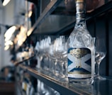 A bottle of Purity Gin is prominently displayed on a bar shelf, surrounded by empty wine glasses. The environment appears to be a polished, modern bar setting with dim lighting reflecting off the glass surfaces.