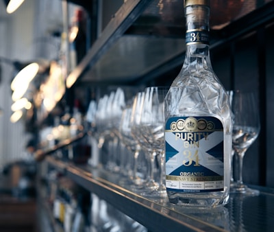 A bottle of Purity Gin is prominently displayed on a bar shelf, surrounded by empty wine glasses. The environment appears to be a polished, modern bar setting with dim lighting reflecting off the glass surfaces.