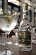 A bottle of Purity Gin 34 is prominently placed on a table, reflecting light with its elegant, faceted design. Next to the bottle is a gin and tonic cocktail garnished with fresh herbs and a slice of lemon, served in a large, rounded glass. The setting appears to be a sophisticated bar or restaurant with blurred backgrounds of glassware and soft lighting, contributing to a refined and classy ambiance.