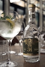 A bottle of Purity Gin 34 is prominently placed on a table, reflecting light with its elegant, faceted design. Next to the bottle is a gin and tonic cocktail garnished with fresh herbs and a slice of lemon, served in a large, rounded glass. The setting appears to be a sophisticated bar or restaurant with blurred backgrounds of glassware and soft lighting, contributing to a refined and classy ambiance.