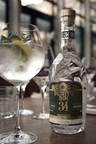 A bottle of Purity Gin 34 is prominently placed on a table, reflecting light with its elegant, faceted design. Next to the bottle is a gin and tonic cocktail garnished with fresh herbs and a slice of lemon, served in a large, rounded glass. The setting appears to be a sophisticated bar or restaurant with blurred backgrounds of glassware and soft lighting, contributing to a refined and classy ambiance.