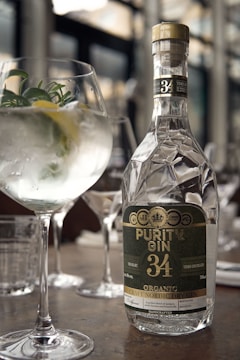 A bottle of Purity Gin 34 is prominently placed on a table, reflecting light with its elegant, faceted design. Next to the bottle is a gin and tonic cocktail garnished with fresh herbs and a slice of lemon, served in a large, rounded glass. The setting appears to be a sophisticated bar or restaurant with blurred backgrounds of glassware and soft lighting, contributing to a refined and classy ambiance.