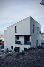 A modern, minimalist white house with a sloped roof located on a rocky terrain. The structure features several windows and a small balcony with a chair and some objects placed on it. Surrounding the building are shrubs and a view of a residential neighborhood with more houses in the background under an overcast sky.