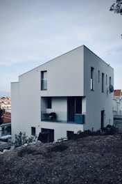 A modern, minimalist white house with a sloped roof located on a rocky terrain. The structure features several windows and a small balcony with a chair and some objects placed on it. Surrounding the building are shrubs and a view of a residential neighborhood with more houses in the background under an overcast sky.