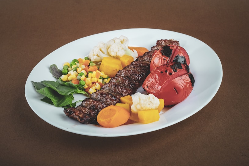 A plate with a variety of colorful cooked vegetables, including roasted red peppers, cauliflower, carrots, peas, corn, and green beans. There are also green leafy vegetables and a grilled skewer of meat.