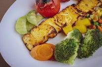 Grilled paneer with a slightly charred surface is served alongside a colorful assortment of vegetables including broccoli, carrots, peas, and corn. A roasted tomato and sliced cucumbers add freshness to the plate.