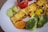 Grilled paneer with a slightly charred surface is served alongside a colorful assortment of vegetables including broccoli, carrots, peas, and corn. A roasted tomato and sliced cucumbers add freshness to the plate.