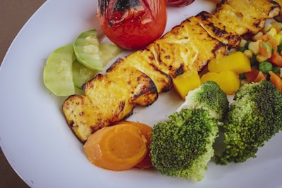 Grilled paneer with a slightly charred surface is served alongside a colorful assortment of vegetables including broccoli, carrots, peas, and corn. A roasted tomato and sliced cucumbers add freshness to the plate.