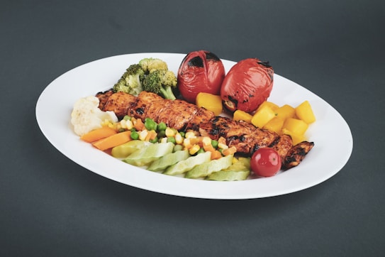 A white plate contains a grilled skewer, surrounded by colorful roasted vegetables including broccoli, tomatoes, carrots, corn, peas, and bell peppers. The dish appears freshly prepared and arranged neatly on a dark background.