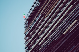 A structure with scaffolding and beams is shown, featuring an Italian flag towards the top against a clear blue sky.