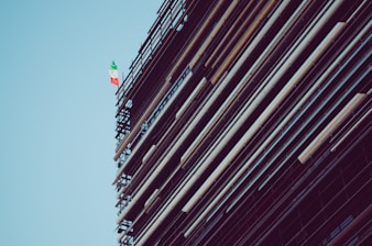 A structure with scaffolding and beams is shown, featuring an Italian flag towards the top against a clear blue sky.