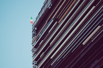 A structure with scaffolding and beams is shown, featuring an Italian flag towards the top against a clear blue sky.
