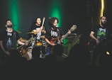 A band is performing on stage with dim lighting and a green spotlight behind them. The musicians, consisting of a drummer, two guitarists, and a bassist, are passionately engaged in their performance. They are wearing dark band-themed t-shirts, and the audience is visible in the foreground as silhouettes.