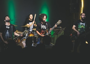 A band is performing on stage with dim lighting and a green spotlight behind them. The musicians, consisting of a drummer, two guitarists, and a bassist, are passionately engaged in their performance. They are wearing dark band-themed t-shirts, and the audience is visible in the foreground as silhouettes.