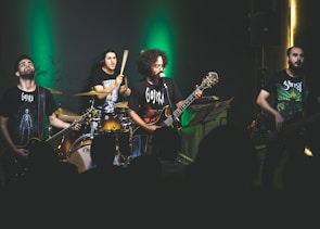 A band is performing on stage with dim lighting and a green spotlight behind them. The musicians, consisting of a drummer, two guitarists, and a bassist, are passionately engaged in their performance. They are wearing dark band-themed t-shirts, and the audience is visible in the foreground as silhouettes.