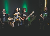 A band is performing on stage with dim lighting and a green spotlight behind them. The musicians, consisting of a drummer, two guitarists, and a bassist, are passionately engaged in their performance. They are wearing dark band-themed t-shirts, and the audience is visible in the foreground as silhouettes.