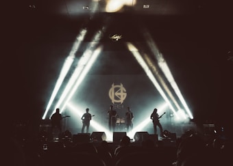 A band of five musicians is performing on a stage with dramatic stage lighting. The lights create a dynamic and visually striking atmosphere, with beams extending from the background towards the audience. The band members are positioned evenly across the stage, each with their instruments. A large emblem or logo is projected on the screen behind them, adding to the visual impact of the scene. The overall setting appears to be a concert or a live performance event.
